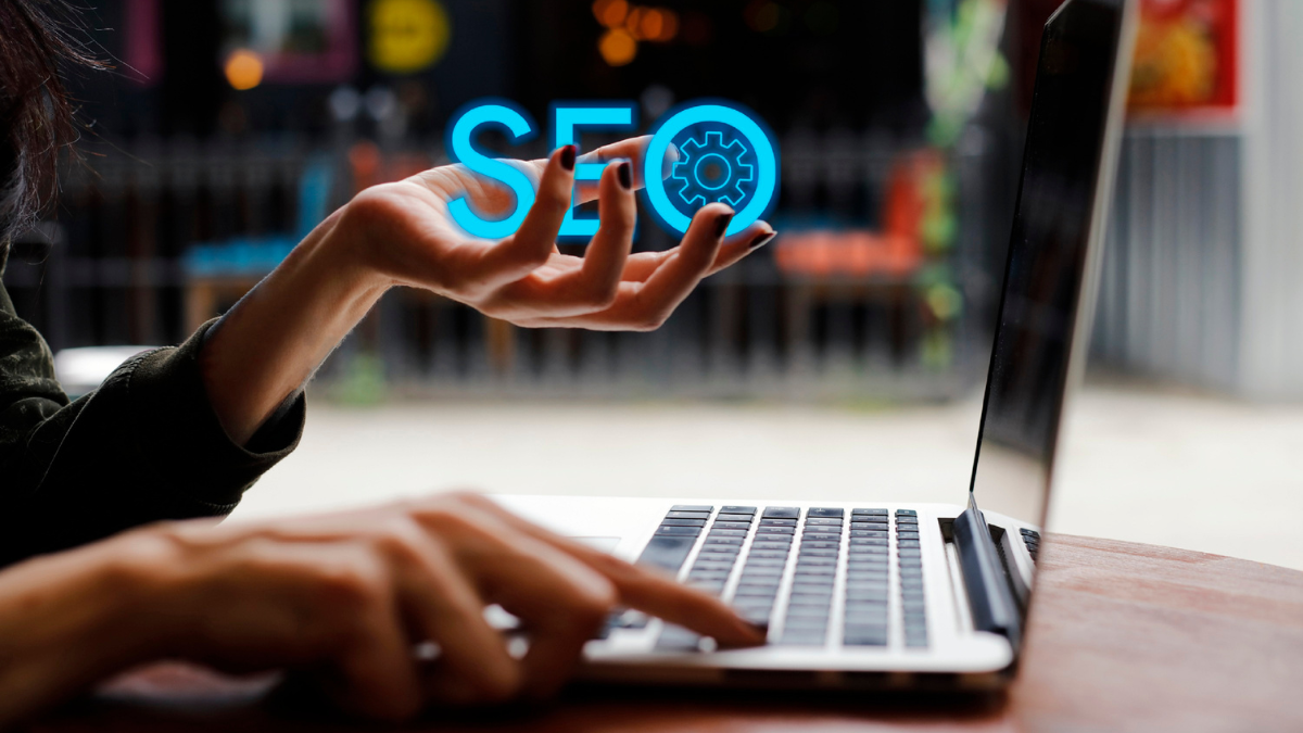 Kobieta pisząca jedną ręką na laptopie, a w drugiej trzymająca napis SEO