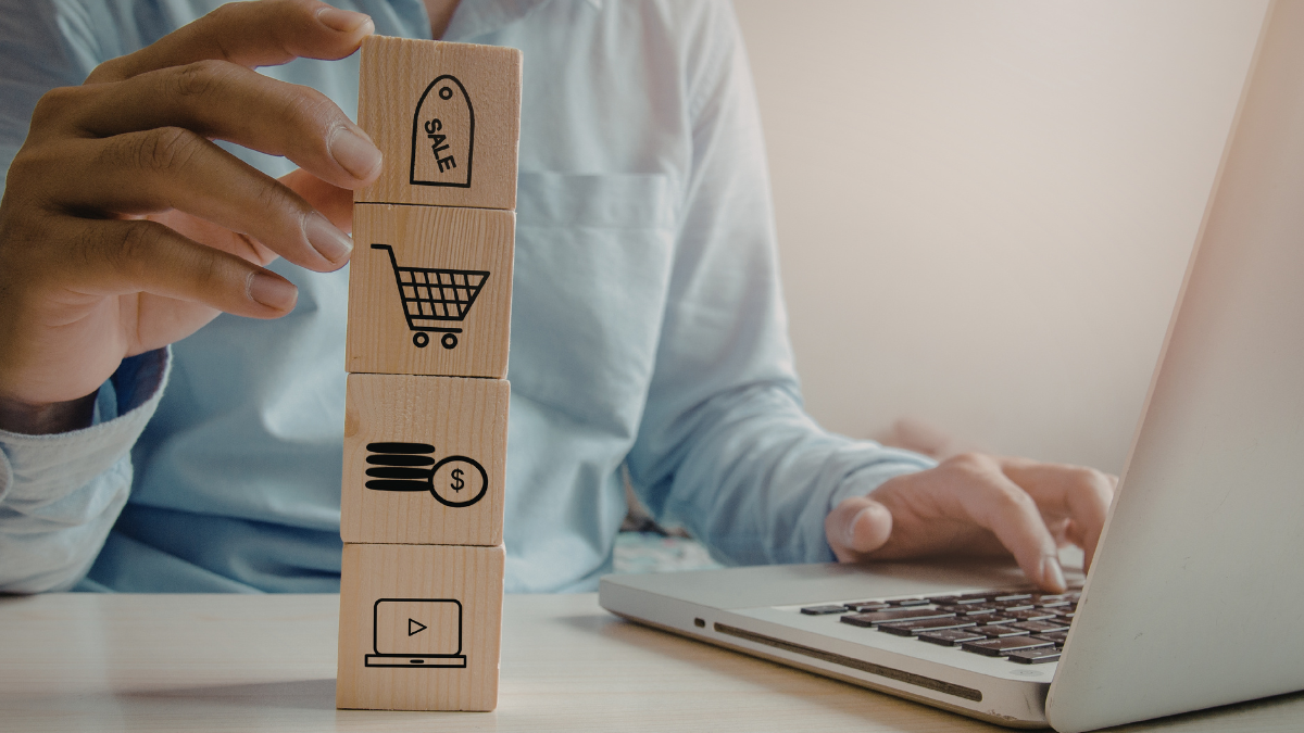 Mężczyzna trzymający jedną ręką wieżę z klocków z symbolami usług e-commerce, drugą pisząc na klawiaturze laptopa